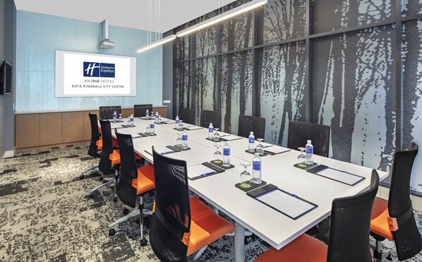 Un salón de conferencias con una mesa larga, sillas, botellas de agua y una pantalla de proyector mostrando un logotipo.