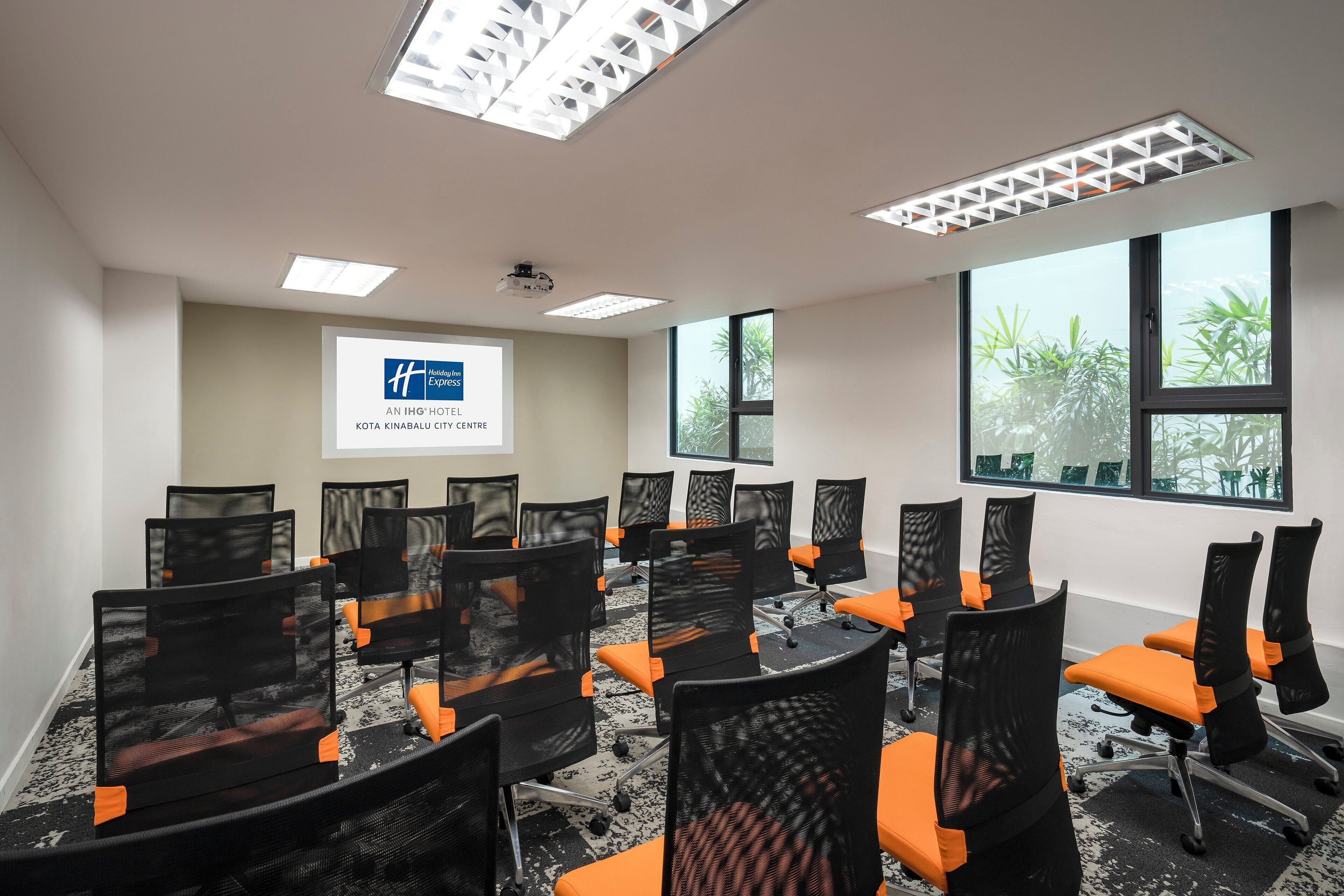 A conference room with black mesh chairs and orange cushions, a projector screen, and large windows with a view of greenery.