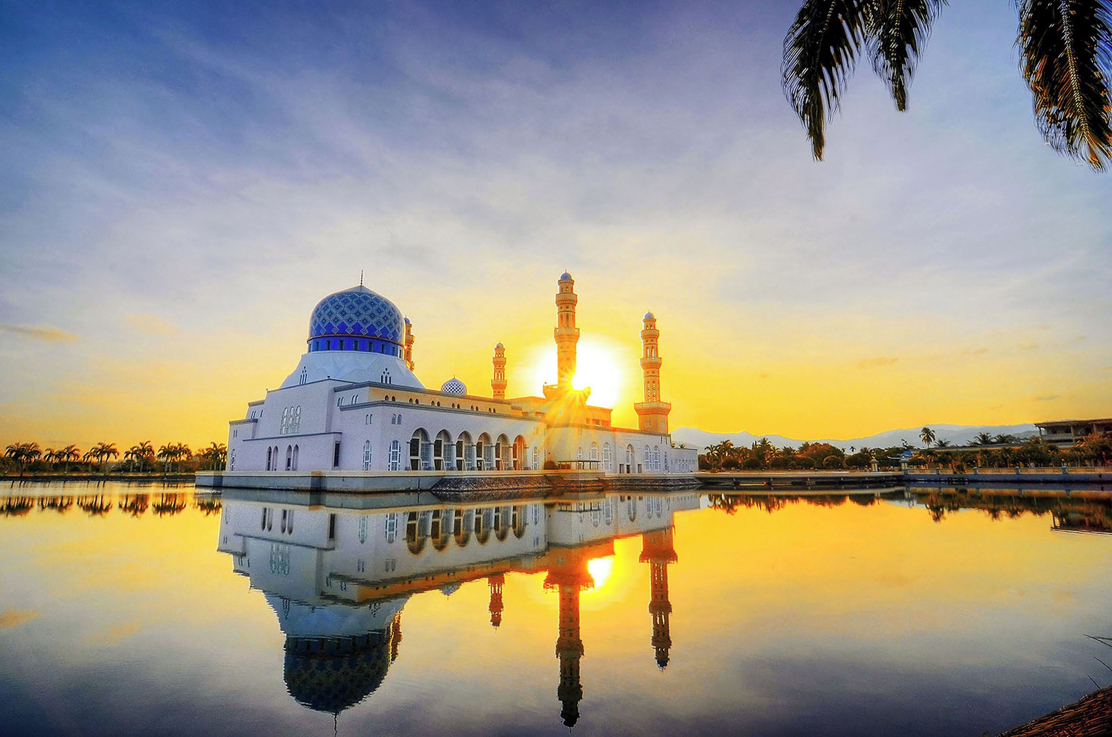 Een moskee met een grote koepel en twee minaretten, weerspiegeld in kalm water tijdens de zonsondergang.