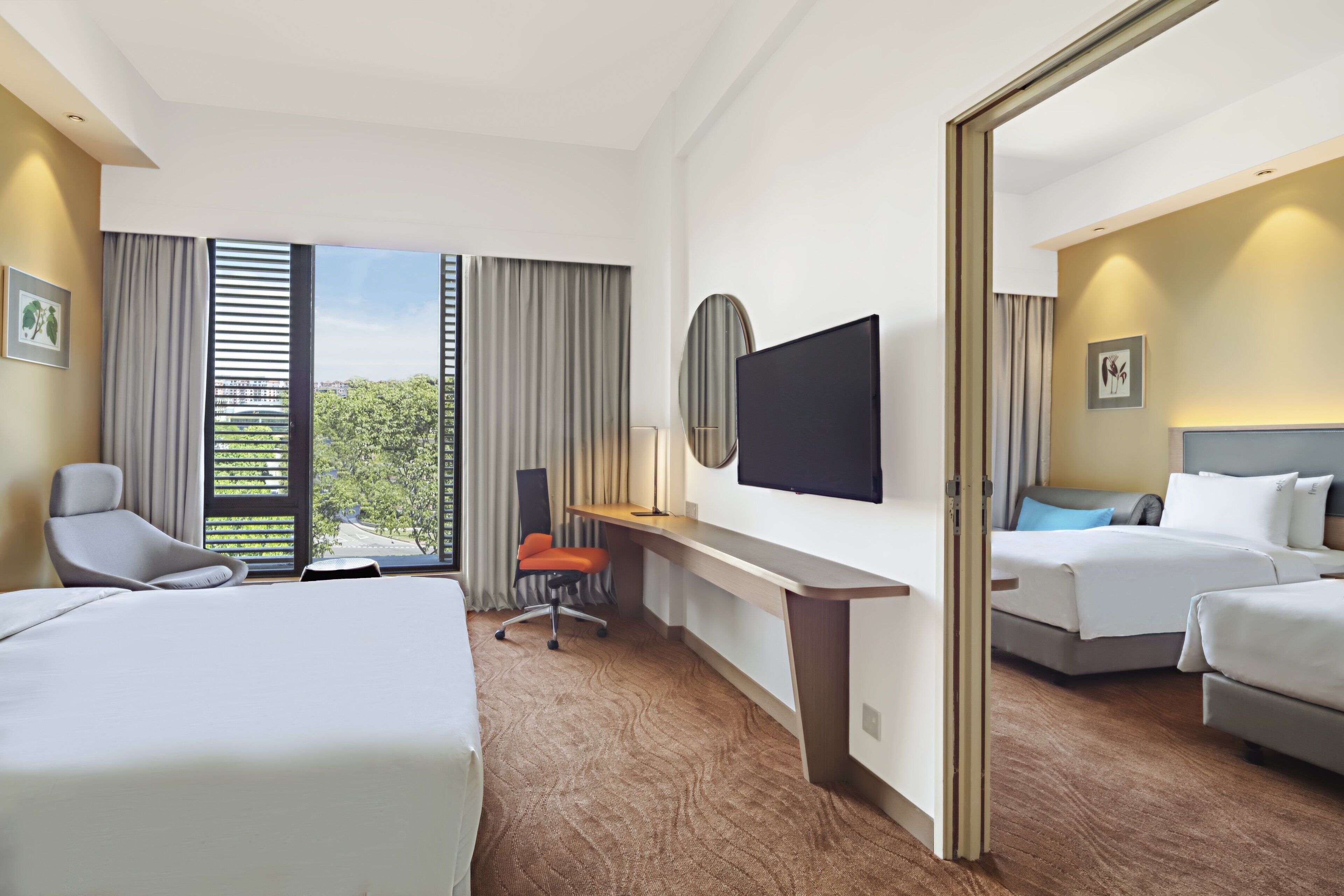 A hotel room with two beds, a desk, a TV, and a view of trees through the windows.