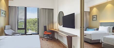 A hotel room with two beds, a desk, a TV, and a view of trees through the windows.