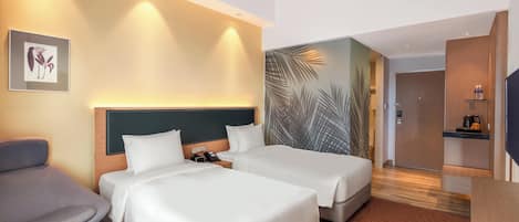 Habitación de hotel con dos camas, un sillón gris, una pequeña mesa redonda y una pared con diseño de hojas de palma.
