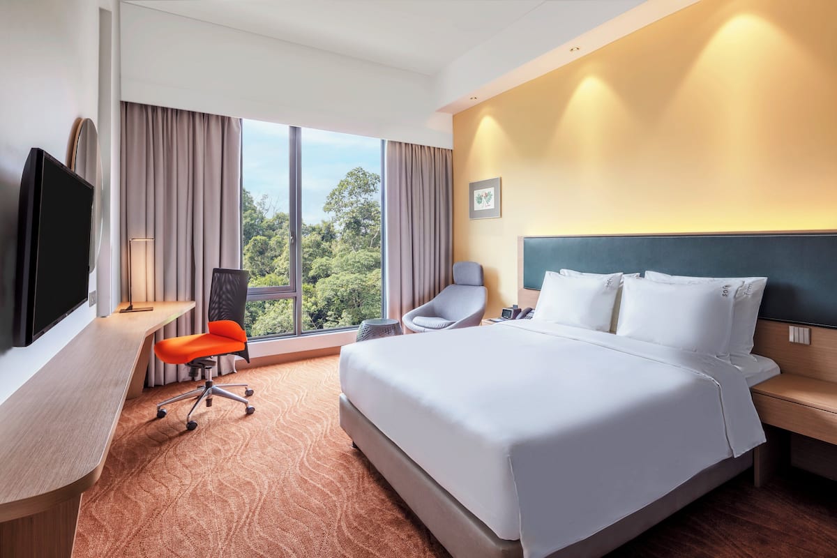 Ein Hotelzimmer mit einem großen Bett, einem Schreibtisch mit Fernseher, einem Sessel und Blick auf Grünanlagen durch das Fenster.