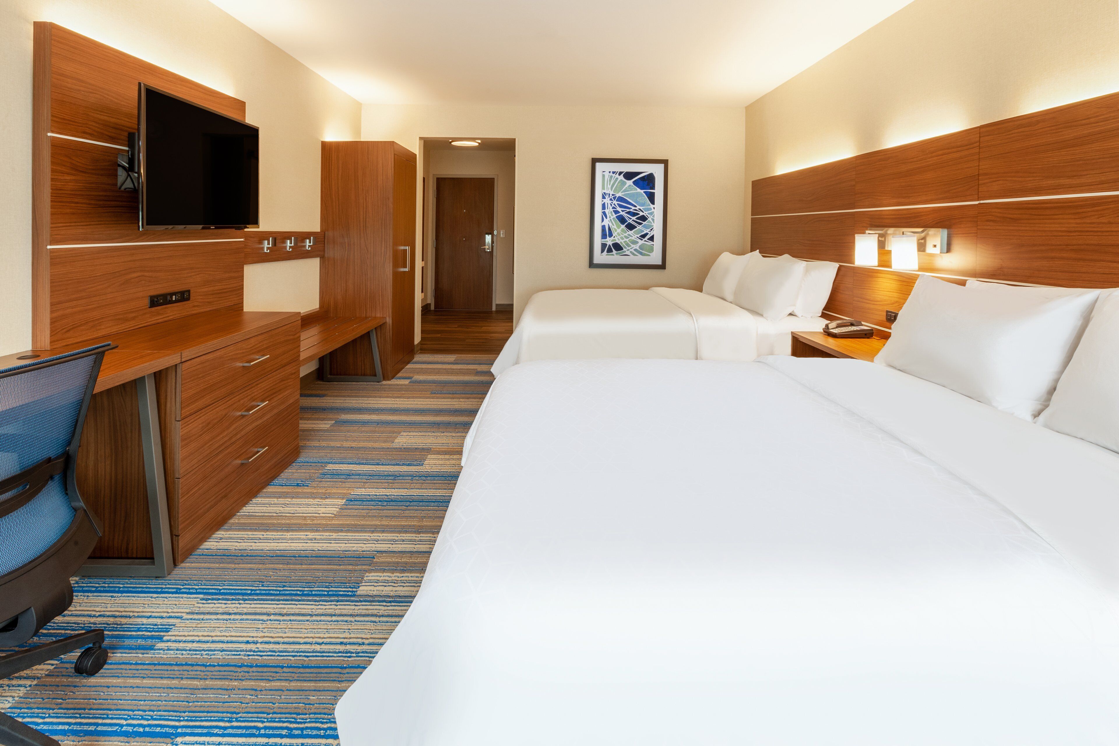Habitación de hotel con una cama grande, un escritorio con silla, un televisor montado en la pared y una obra de arte enmarcadas en la pared.