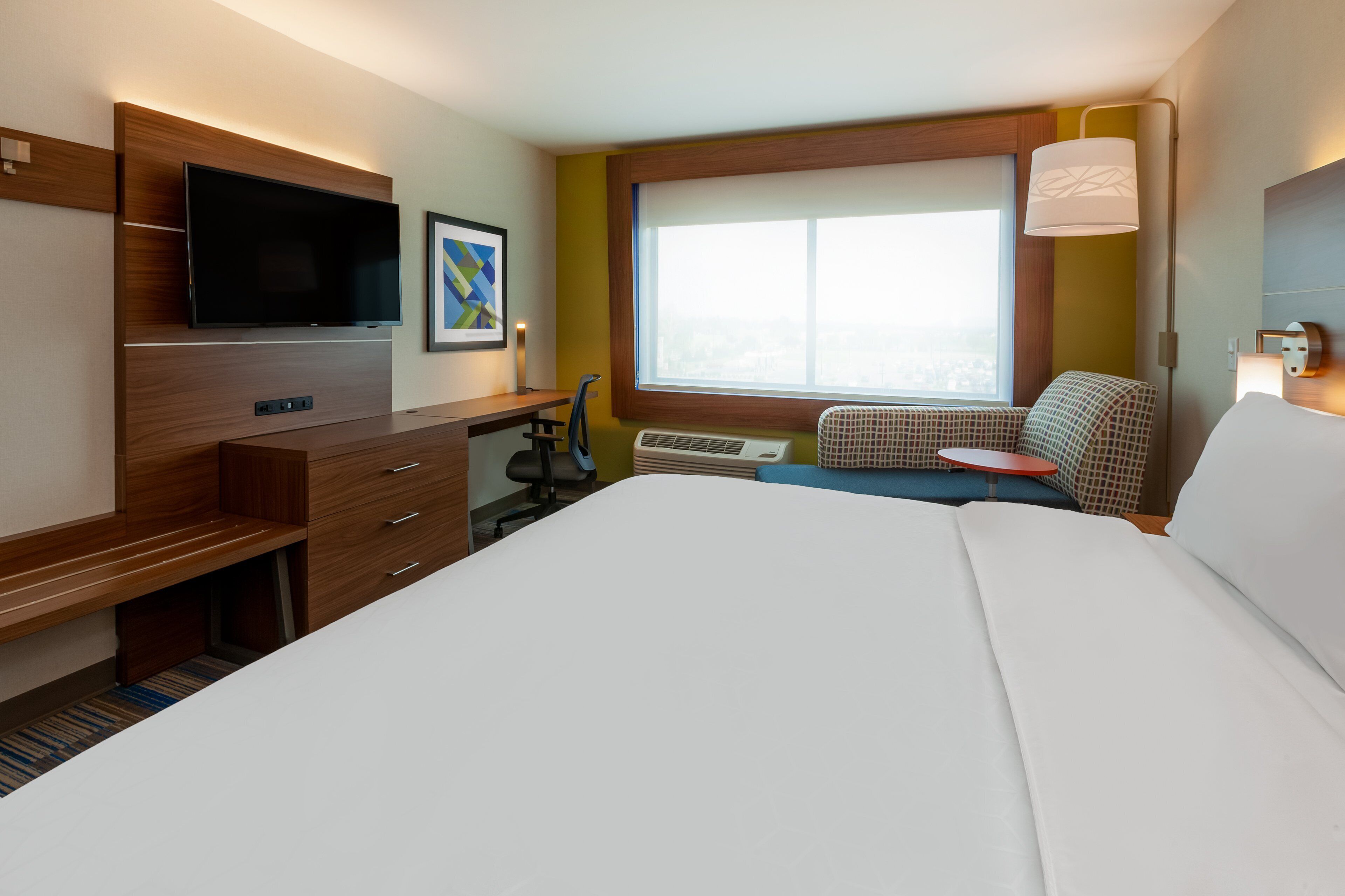 A hotel room with a large bed, a desk with a flat-screen TV, a window with a view, and a seating area with a lamp.