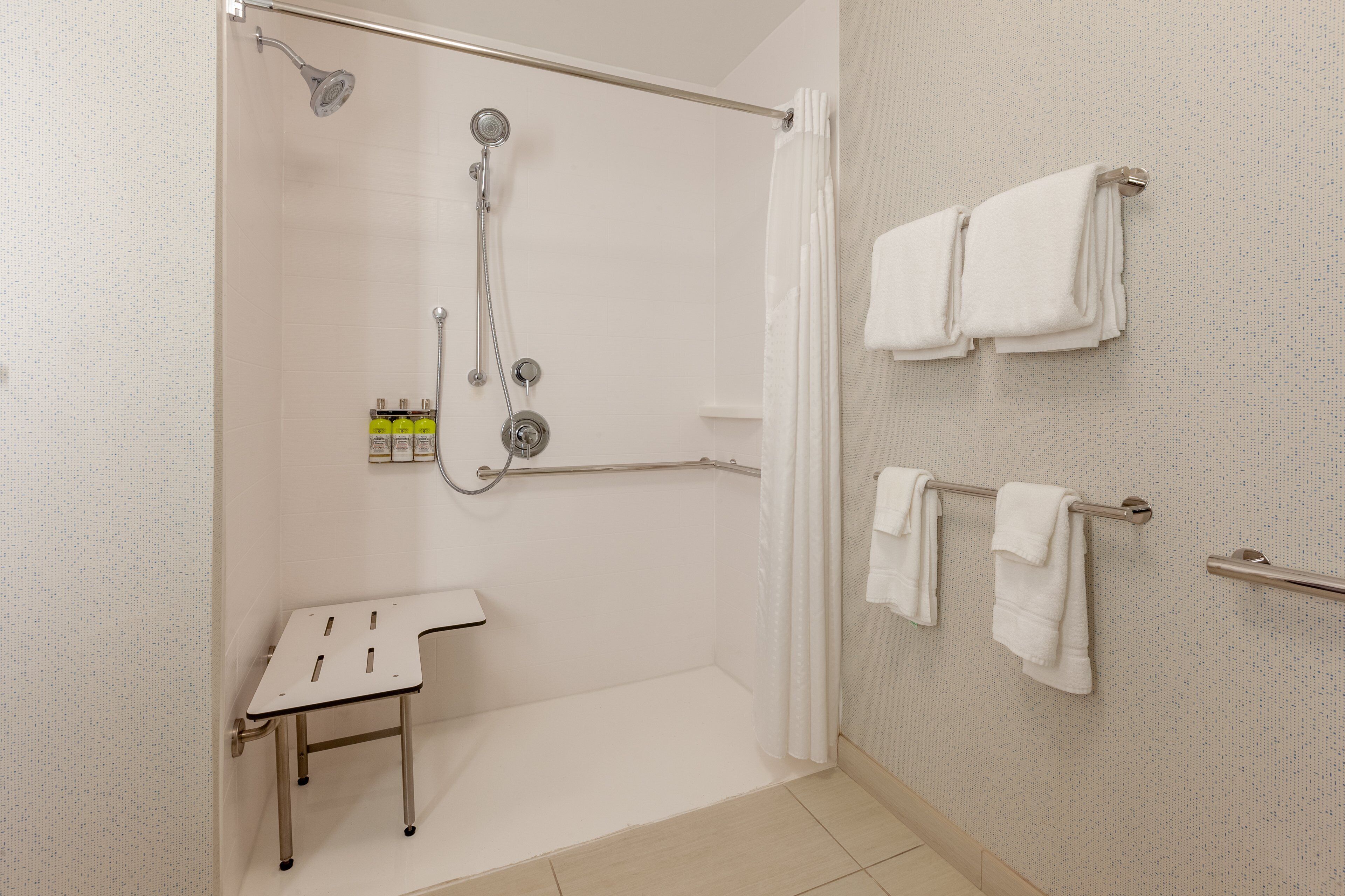 Casa de banho moderna com chuveiro amplo, lavatório branco e várias toalhas penduradas num porta-toalhas.