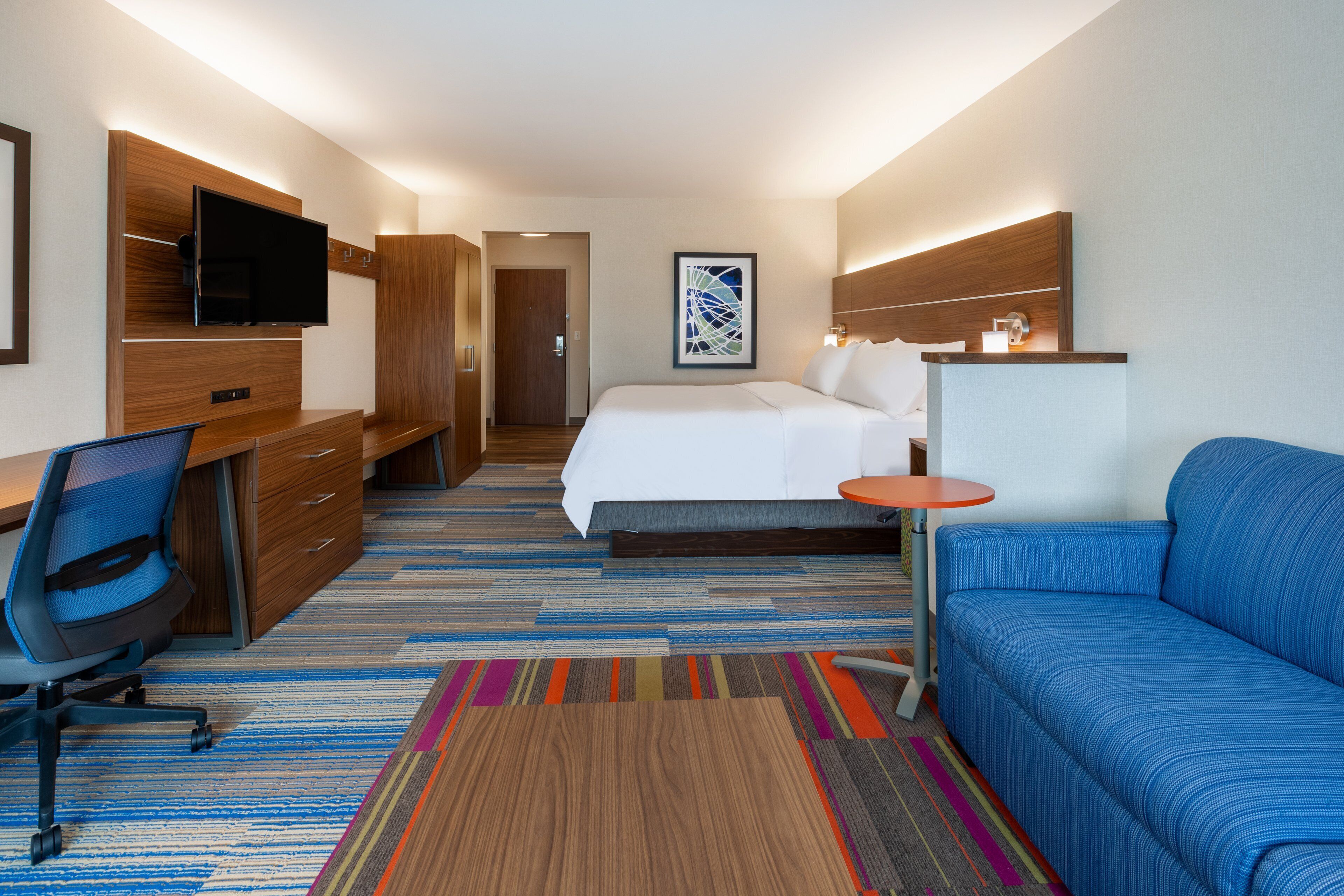 A hotel room with a bed, a desk with a chair, a TV mounted on a wooden panel, a colorful striped rug, and a blue sofa.