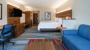A hotel room with a bed, a desk with a chair, a TV mounted on a wooden panel, a colorful striped rug, and a blue sofa.
