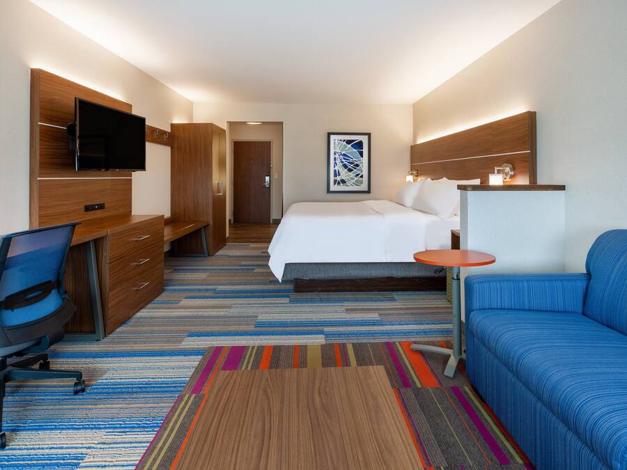 A hotel room with a bed, a desk with a chair, a TV mounted on a wooden panel, a colorful striped rug, and a blue sofa.