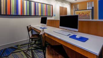 A reception area with a long desk, two computer monitors, a keyboard, and a mouse. There are two chairs, a carpeted floor, and a colorful wall art piece.