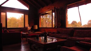 Ein gemütliches Blockhaus-Interieur mit Holzboden, einer Couch, einem Couchtisch und großen Fenstern mit Blick auf Bäume.