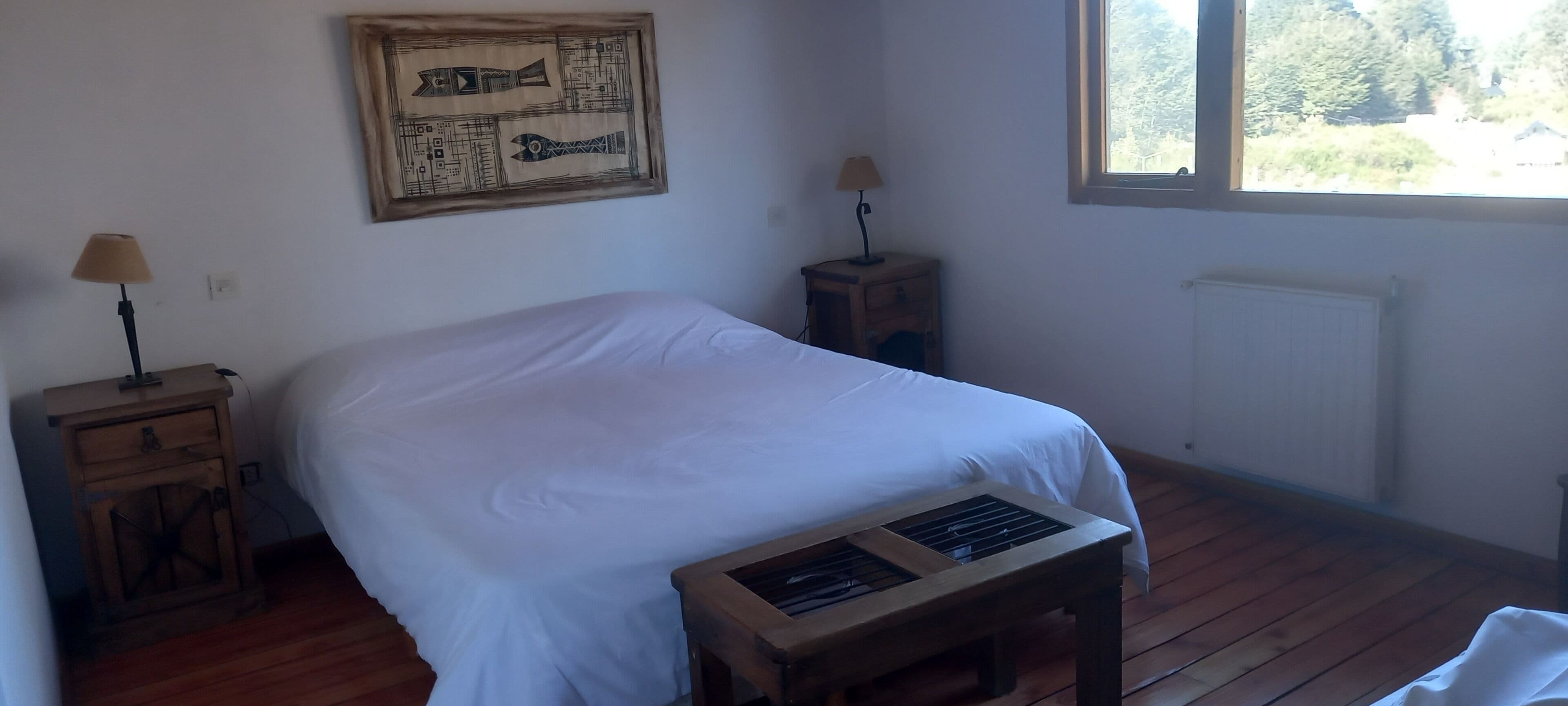 Une chambre à coucher avec un lit, des tables de chevet en bois, une table, une fenêtre et un radiateur.
