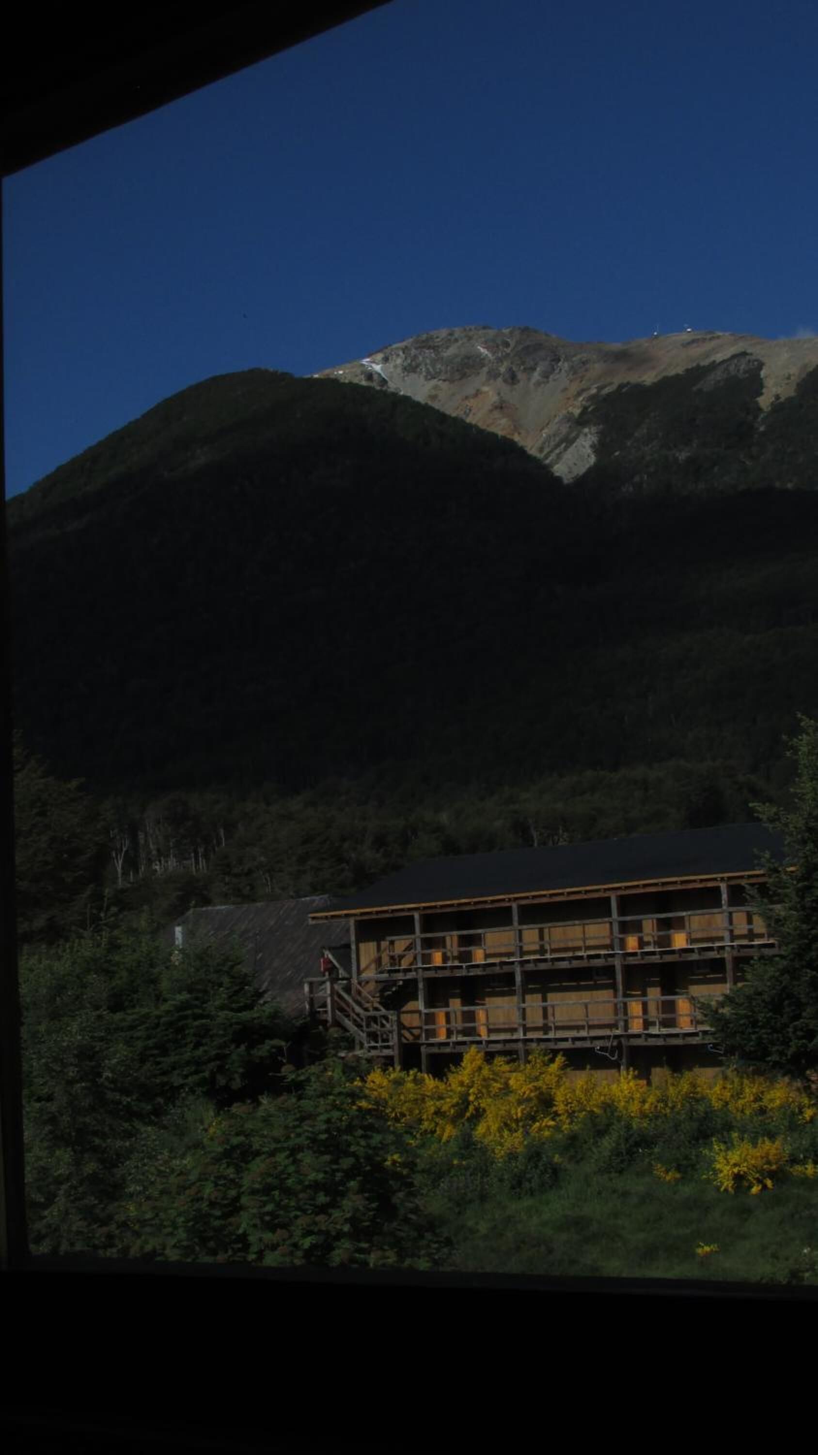 Une cabane de montagne avec une façade en bois, un escalier menant à l’entrée et une vue sur une chaîne de montagnes en arrière-plan.