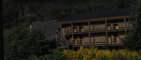 Une cabane de montagne avec une façade en bois, un escalier menant à l’entrée et une vue sur une chaîne de montagnes en arrière-plan.