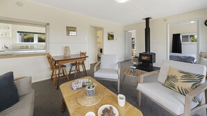 A cozy living room with a fireplace, a dining area with wooden furniture, and a view of the outside through a window. - The Green Trout - Paraparaumu Beach Holiday Home (Paraparaumu Beach)
