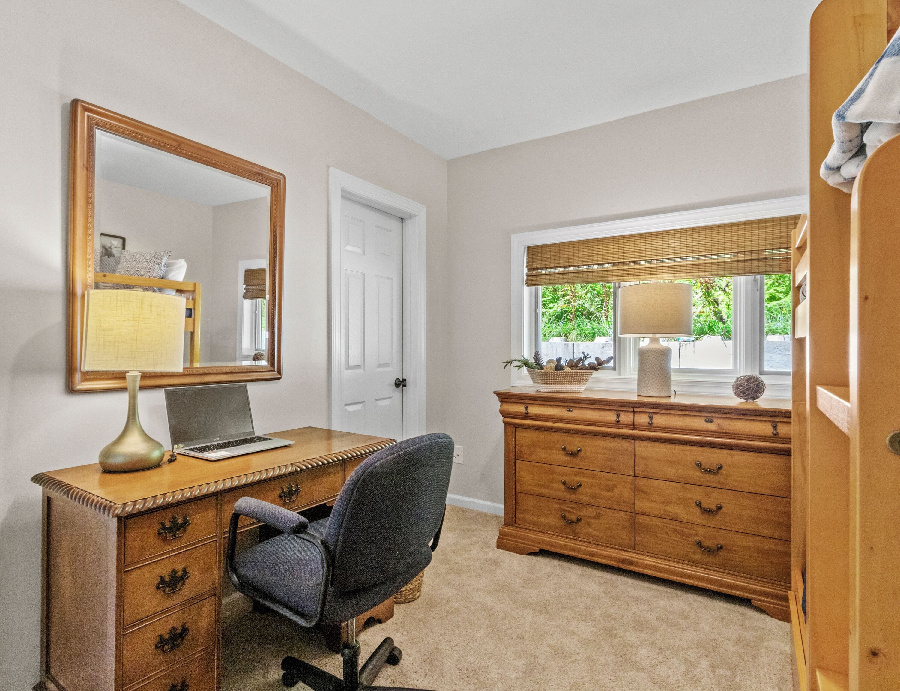 Une pièce comprenant un bureau, une chaise, une commode, un miroir et une fenêtre avec des stores.