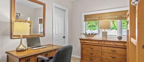 Une pièce comprenant un bureau, une chaise, une commode, un miroir et une fenêtre avec des stores.