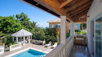 Un espace piscine comprenant des chaises longues, un kiosque et une terrasse couverte offrant une vue sur une maison et la verdure environnante.