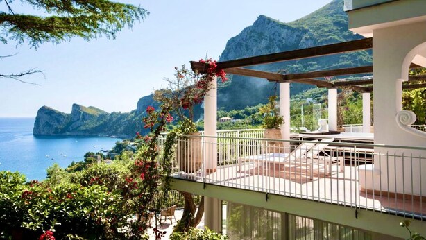 Un balcon offrant une vue sur la mer et les montagnes, agrémenté d’une pergola ornée de fleurs et de mobilier d’extérieur.