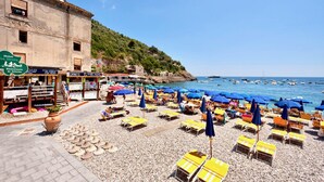 Une plage avec de nombreux transats et parasols, un bâtiment sur lequel on peut voir une enseigne indiquant un restaurant, et des bateaux sur l’eau.