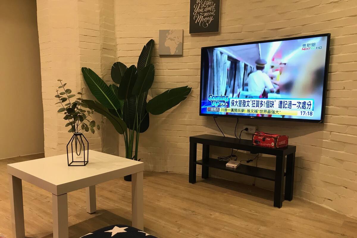 A living room with a television mounted on the wall, a small white table with a vase of flowers, a large green plant, and a black entertainment unit with a red speaker.
