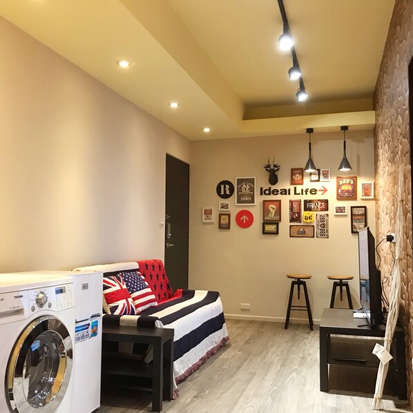 Una lavandería con lavadora, una cama con manta de la bandera estadounidense y una pared con varios objetos enmarcados y un cartel que dice “Ideal Life”.