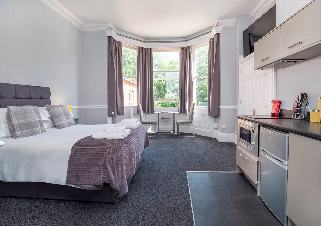 A modern hotel room with a large bed, a small dining table, and a kitchenette.