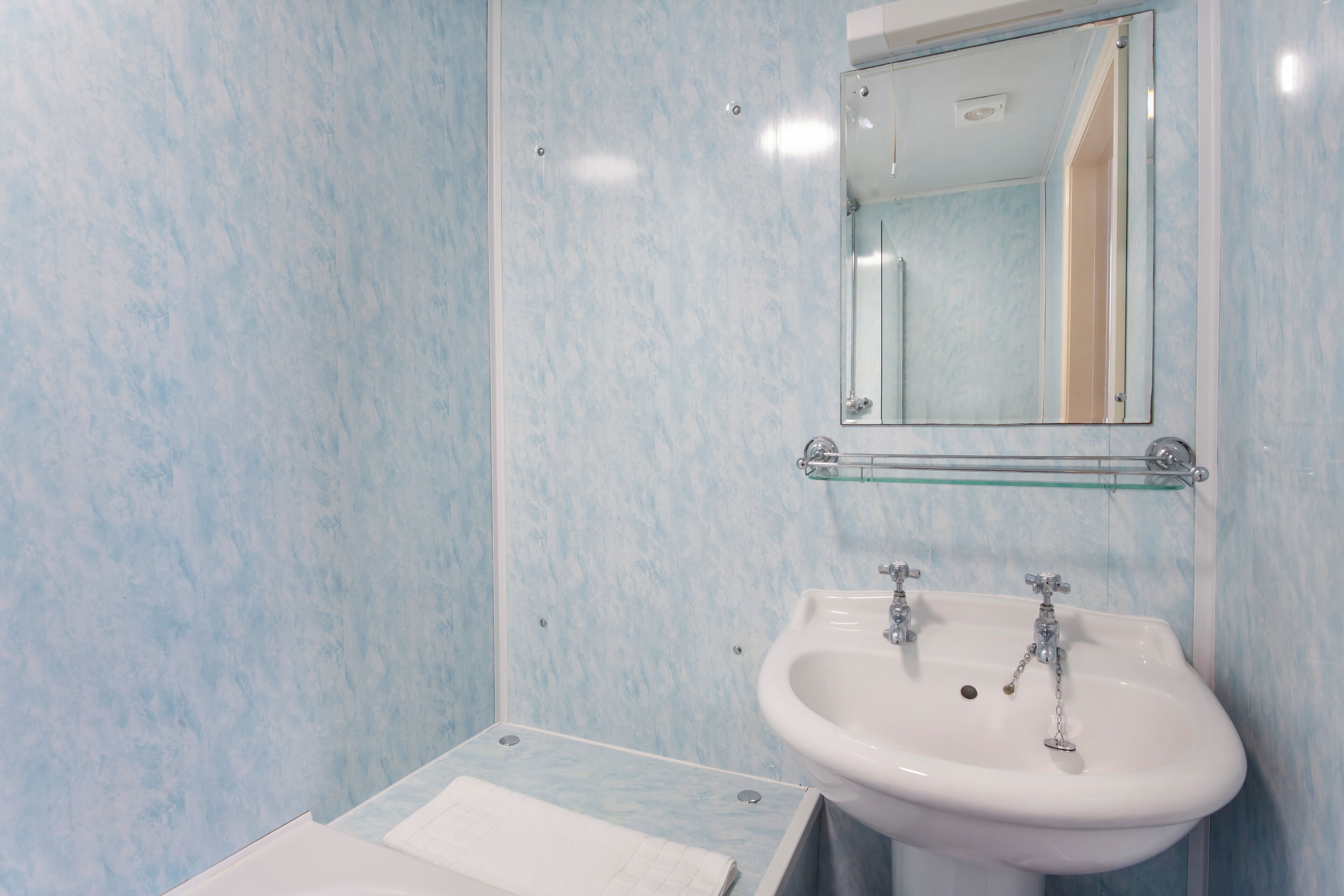 A bathroom with a white sink, two faucets, a glass shelf above the sink, and a mirror.