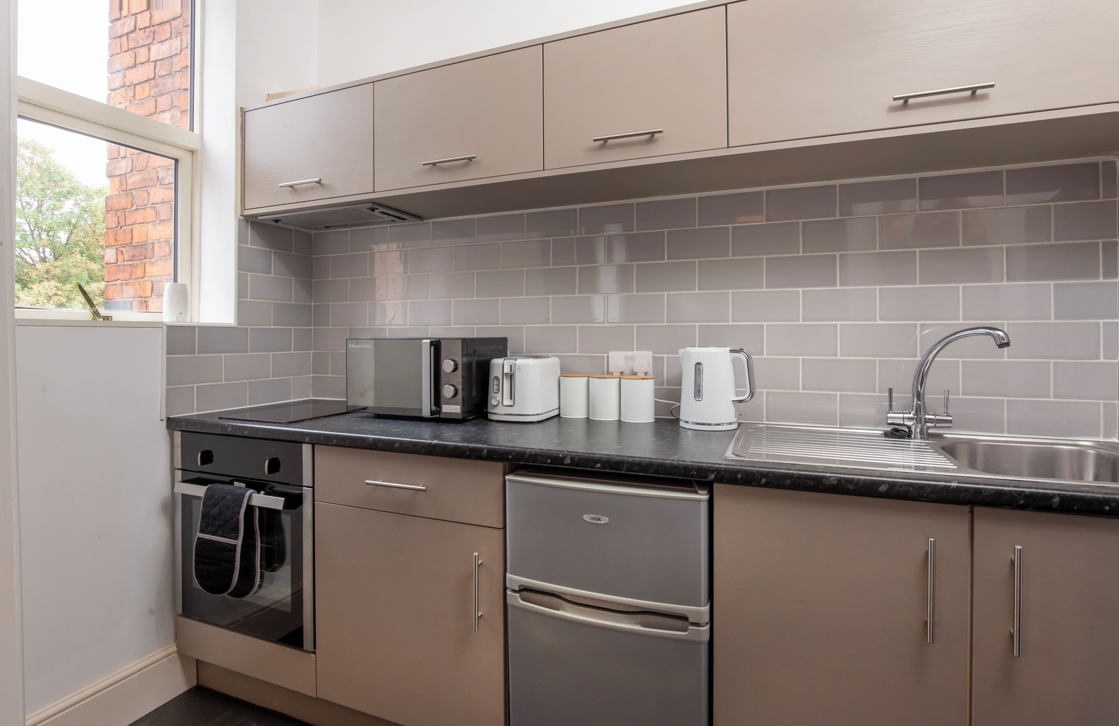 A modern kitchen with grey cabinets, a stainless steel refrigerator, a built-in oven, a sink, and a window with a view of a brick building.