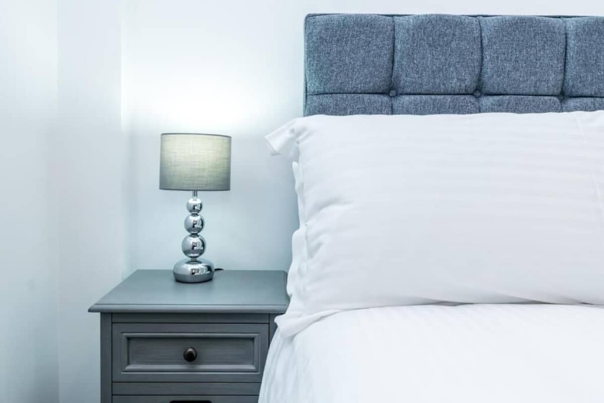 A neatly made bed with a grey headboard and a bedside table with a lamp.