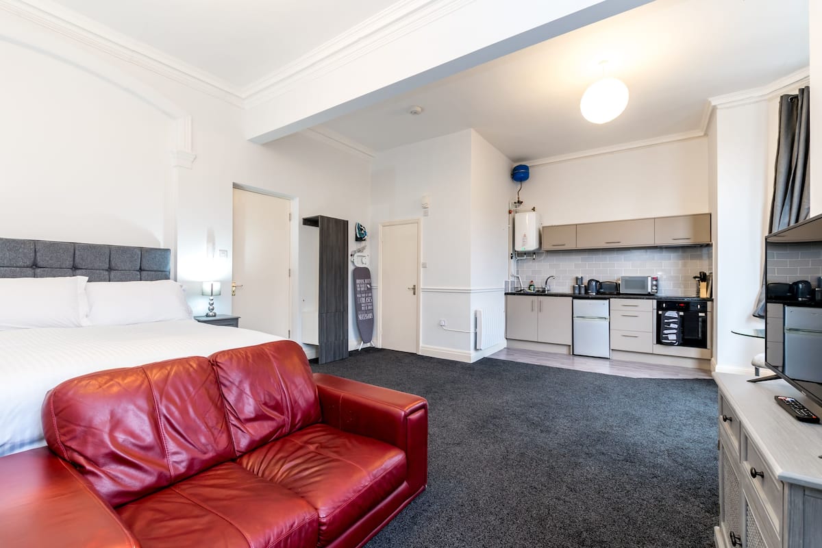 A hotel room with a red leather sofa, a bed with white linens, a kitchenette, and a bathroom with a towel hanging on the door.