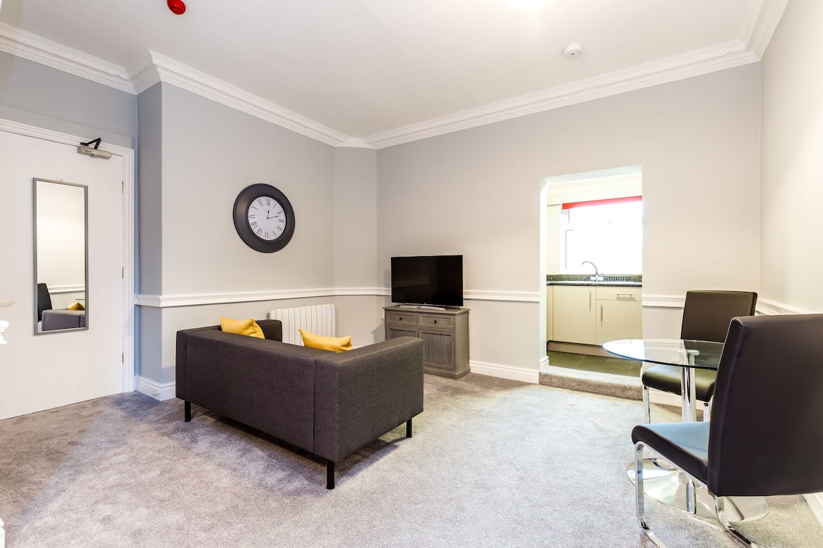 A modern living room with a grey sofa, a flat-screen TV, and a dining area with a glass table and chairs.