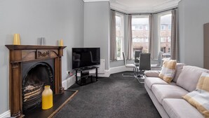 A living room with a fireplace, a television, a sofa, and a view of buildings through large windows. - Sycamore Apartments (York)