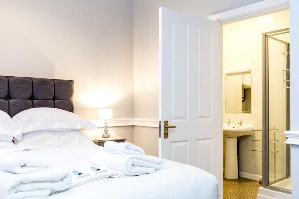 A hotel room with a bed, a bedside table with a lamp, a bathroom with a sink and mirror, and a door leading to another room.
