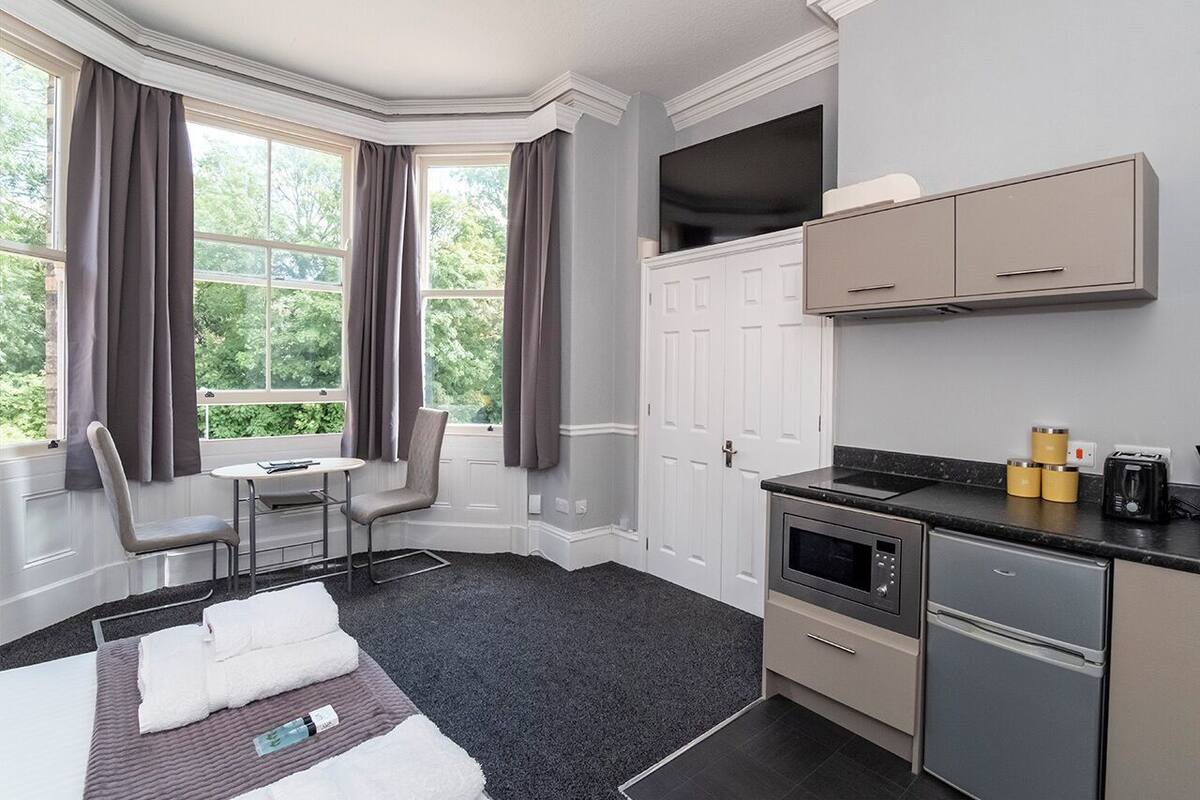 A hotel room with a bed, a desk, a chair, a small table, a window with curtains, a kitchenette with a stove, a microwave, a refrigerator, and cabinets.