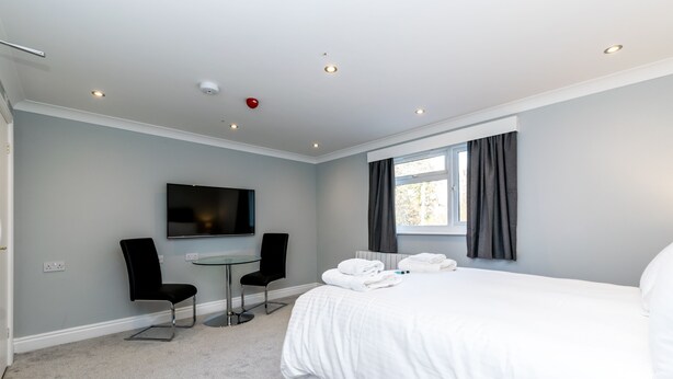 A modern hotel room with a large bed, a flat-screen TV, two chairs, and a small table. - Sycamore Apartments (York)