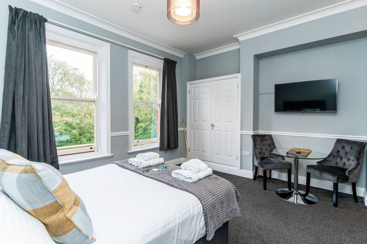 A hotel room with a bed, two chairs, a small table, a TV, and a window with curtains.