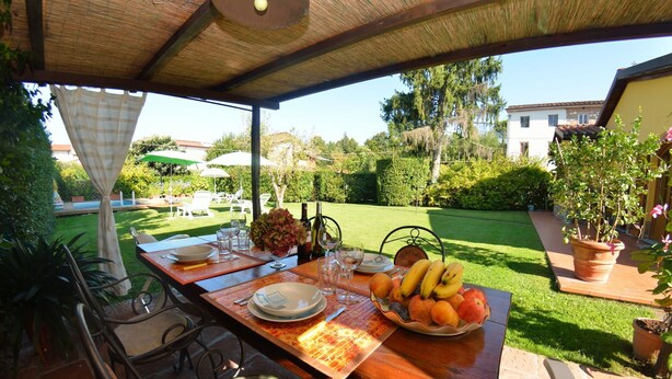 An outdoor dining area with a table set for two, surrounded by chairs, a pool, and a garden.