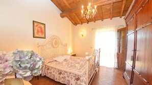 A bedroom with a wooden ceiling, a bed with a floral bedspread, a nightstand, a wall-mounted lamp, and a wooden wardrobe.