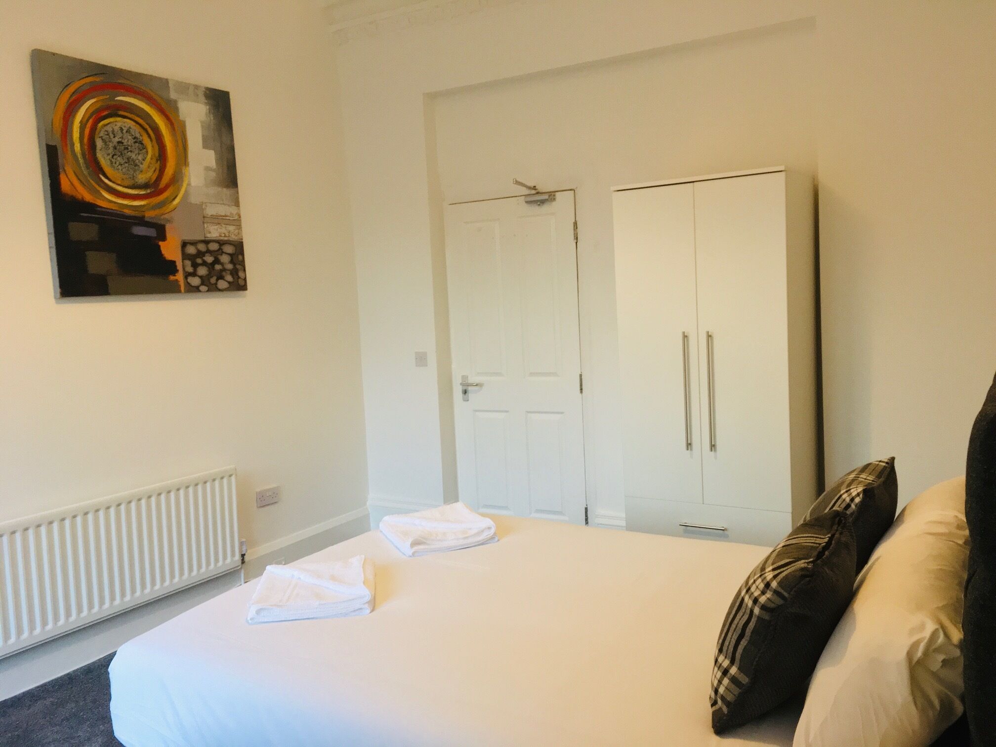 A hotel room with a bed, white towels, a radiator, a wardrobe, and a painting on the wall.