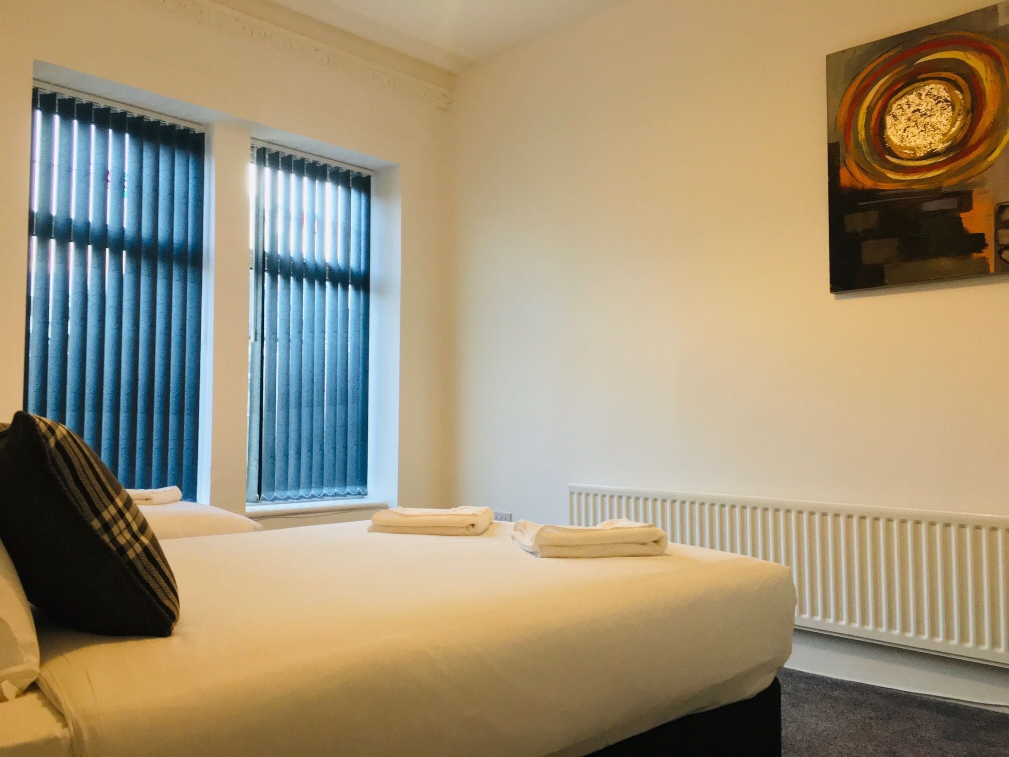 A hotel room with a bed, two windows with blinds, a radiator, and a painting on the wall.