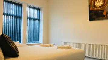 A hotel room with a bed, two windows with blinds, a radiator, and a painting on the wall.