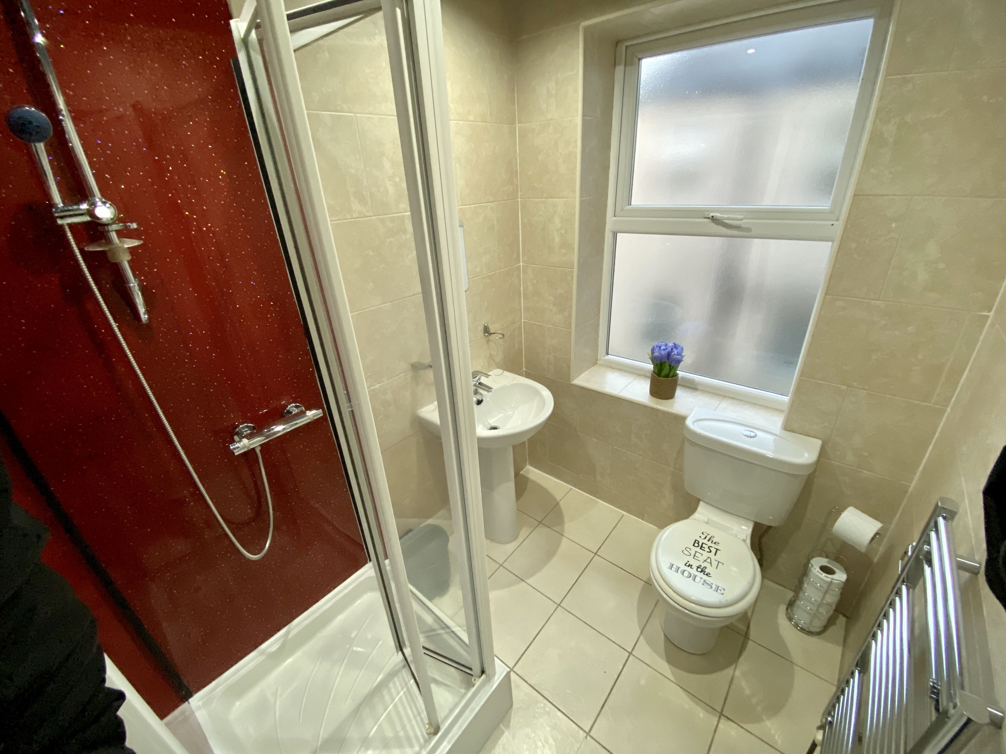 A bathroom with a glass shower enclosure, a sink, a toilet with a decorative seat, and a window with a view.
