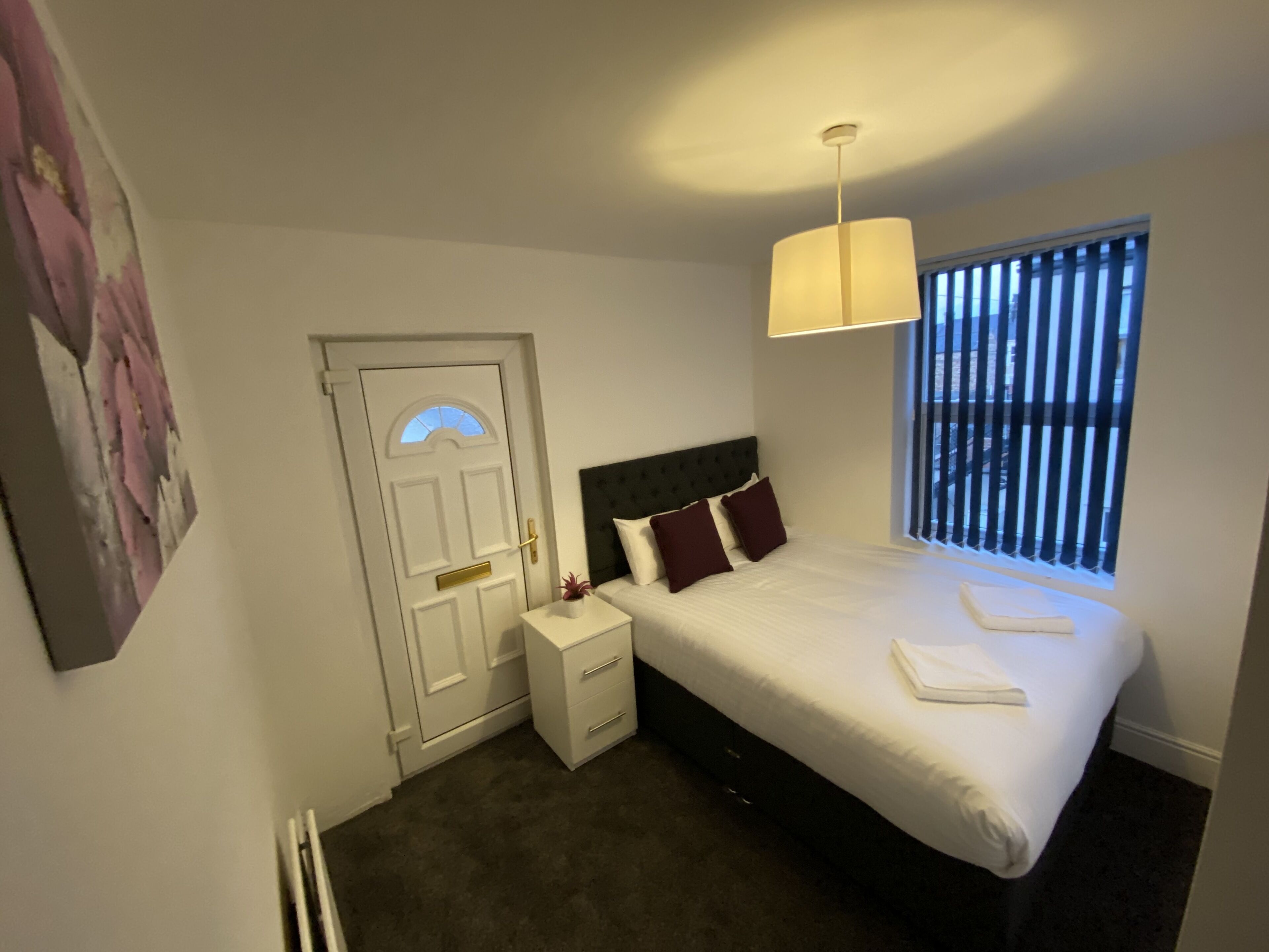 A bedroom with a bed, bedside table, lamp, window, and a door.