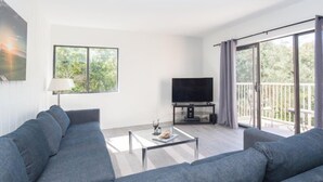 A modern living room with a grey sofa set, a flat-screen TV on a stand, a glass coffee table, and a balcony with a view of greenery. - 2077 Beachwood (Fernandina Beach)