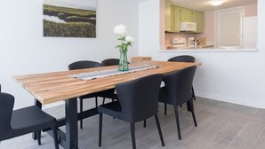 A dining area with a wooden table and black chairs, a small vase with white flowers, and a kitchen in the background. - 2077 Beachwood (Fernandina Beach)