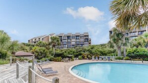 A pool area with lounge chairs, a wooden deck, and a building with multiple balconies in the background. - 1149 Beachwalker (Fernandina Beach)