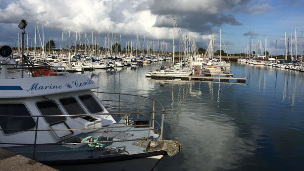 Un puerto deportivo con varias embarcaciones amarradas, un barco llamado 'Marine Camille' en primer plano, y un cielo despejado con nubes.