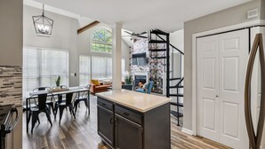 A modern kitchen with a central island, dining area with chairs, a staircase, and a large window with blinds.