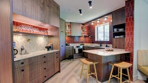 A modern kitchen with wooden cabinetry, a central island with stools, a built-in oven, and a window providing natural light. - Designer 4BR, 3 Bath Townhouse w Mtn Views, Hot Tub, Parking for 2 vehicles (Whistler)